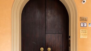 Puerta doble de madera oscura con manija de bronce, ubicada en un vano de arco, sobre una pared amarilla con una cámara de seguridad y un letrero que indica el número de habitación 2.