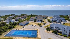 An aerial view of a residential area with houses, a tennis court, and a large body of water in the background. - NanDUCKet on the bay in DUCK, NC! (Kitty Hawk)
