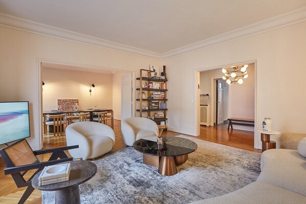 A modern living room with a sofa, armchair, and a coffee table. There is a bookshelf and a dining area in the background. - Apartment Arc de Triomphe/ Champs Elysee (Paris)