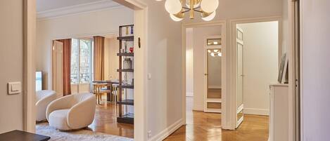 Un vaste espace de vie agrémenté d’un lustre moderne, un coin repas avec une table et des chaises, et un couloir menant à une chambre.