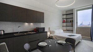 A modern hotel room with a kitchenette, a dining area, a bed, and a balcony with a view of the city.