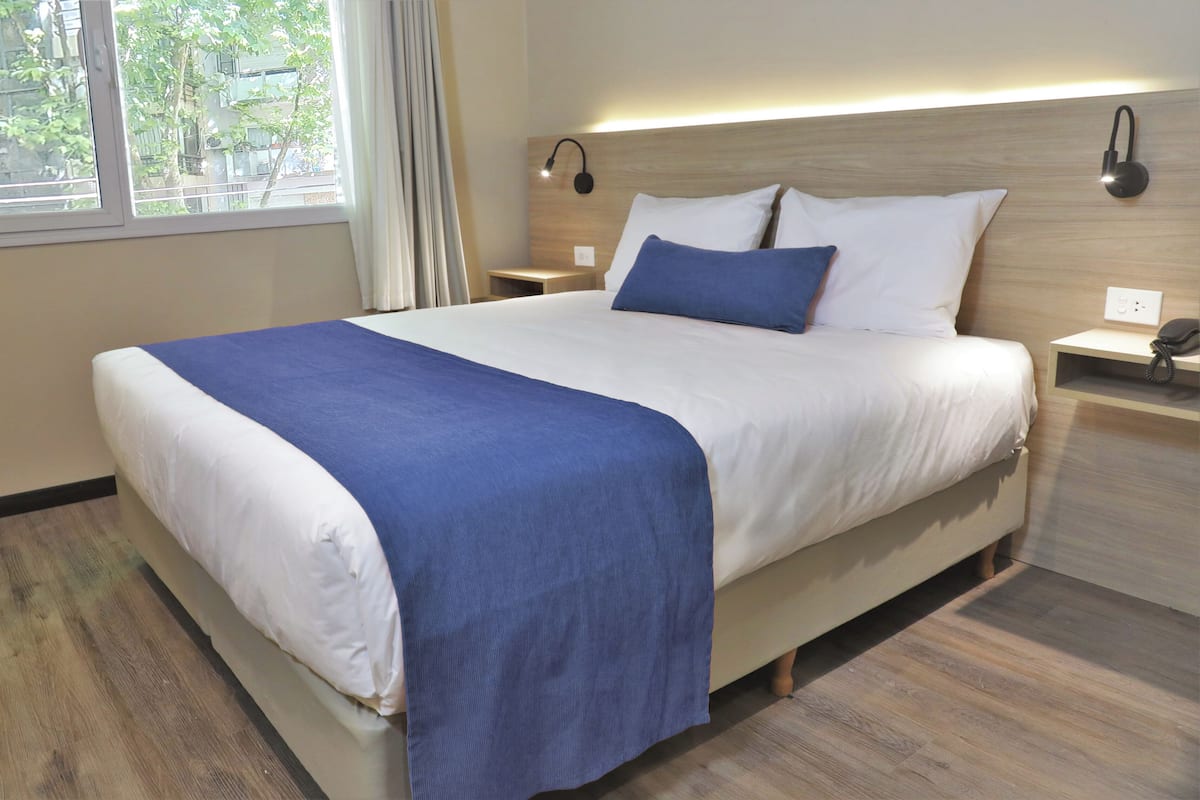 A hotel room with a large bed, a nightstand, a wall-mounted lamp, and a window with curtains.