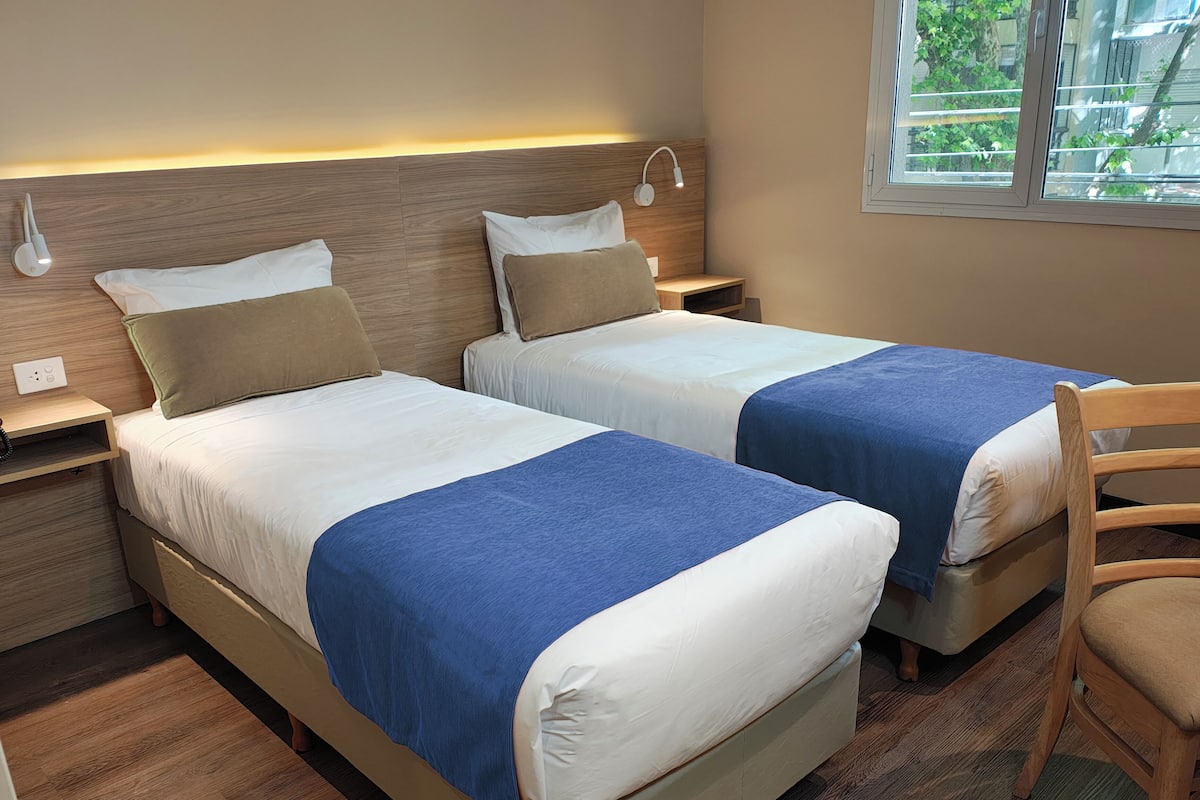 A hotel room with two beds, each with a blue blanket, wooden headboards, and bedside lamps.