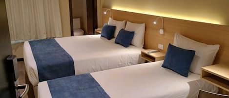 A hotel room with two beds, each with a blue bedspread and white pillows. There is a bedside table with a lamp on each side of the beds.