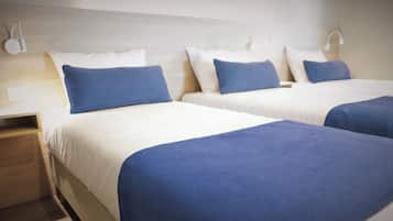 Camera d'albergo con due letti, ognuno con lenzuola bianche e cuscini blu. C'è un comodino in legno con una lampada e una presa elettrica.