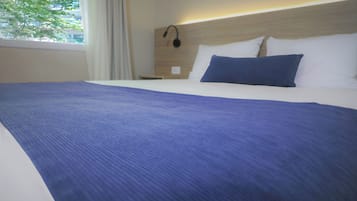 Habitación de hotel con una cama grande, ropa de cama blanca, una manta azul y vista a los árboles por la ventana.