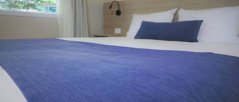 A hotel room with a large bed, white bedding, a blue blanket, and a view of trees through the window.