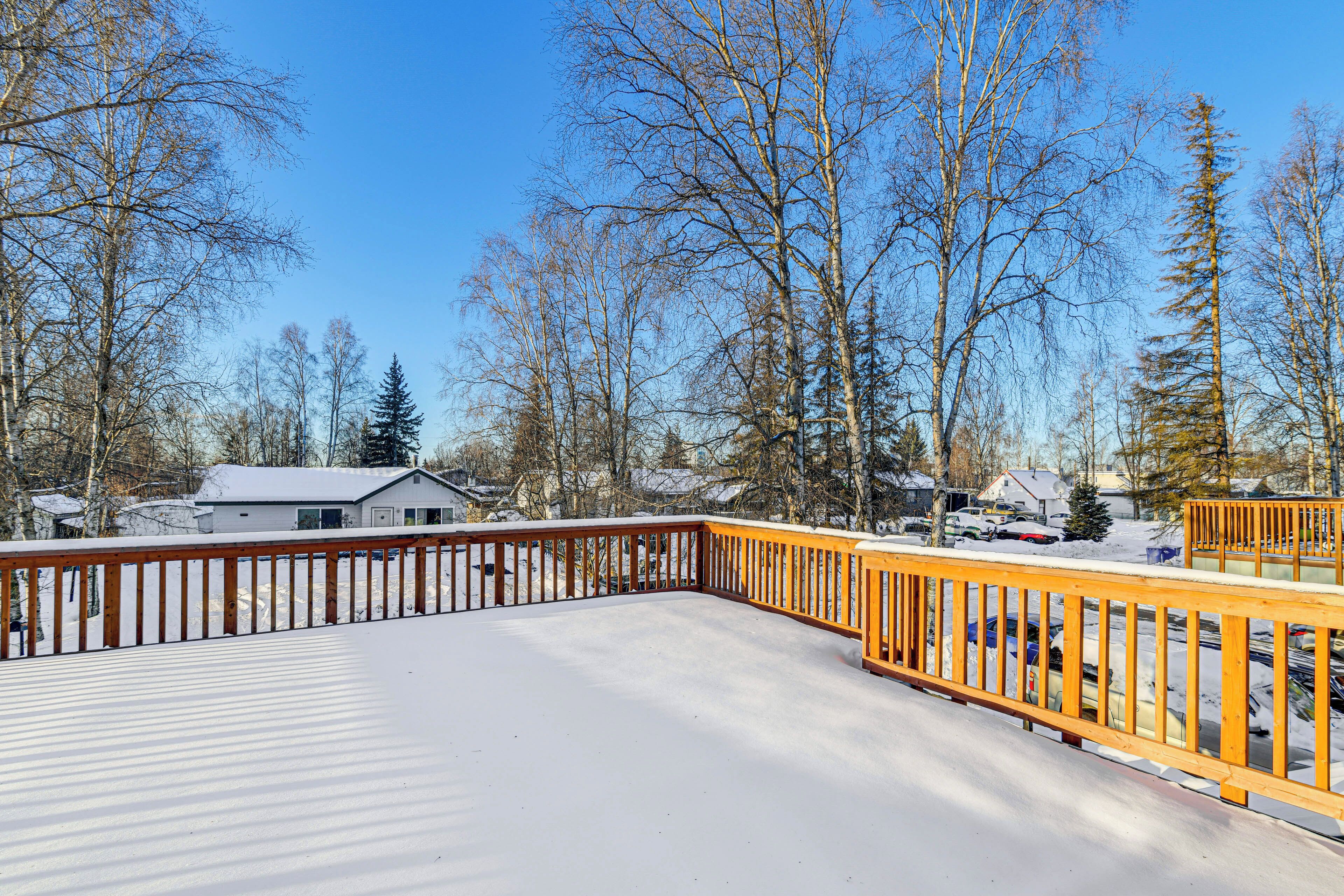 A snow-covered deck with a wooden railing, surrounded by leafless trees and houses in the background.