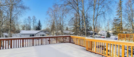 A snow-covered deck with a wooden railing, surrounded by leafless trees and houses in the background.