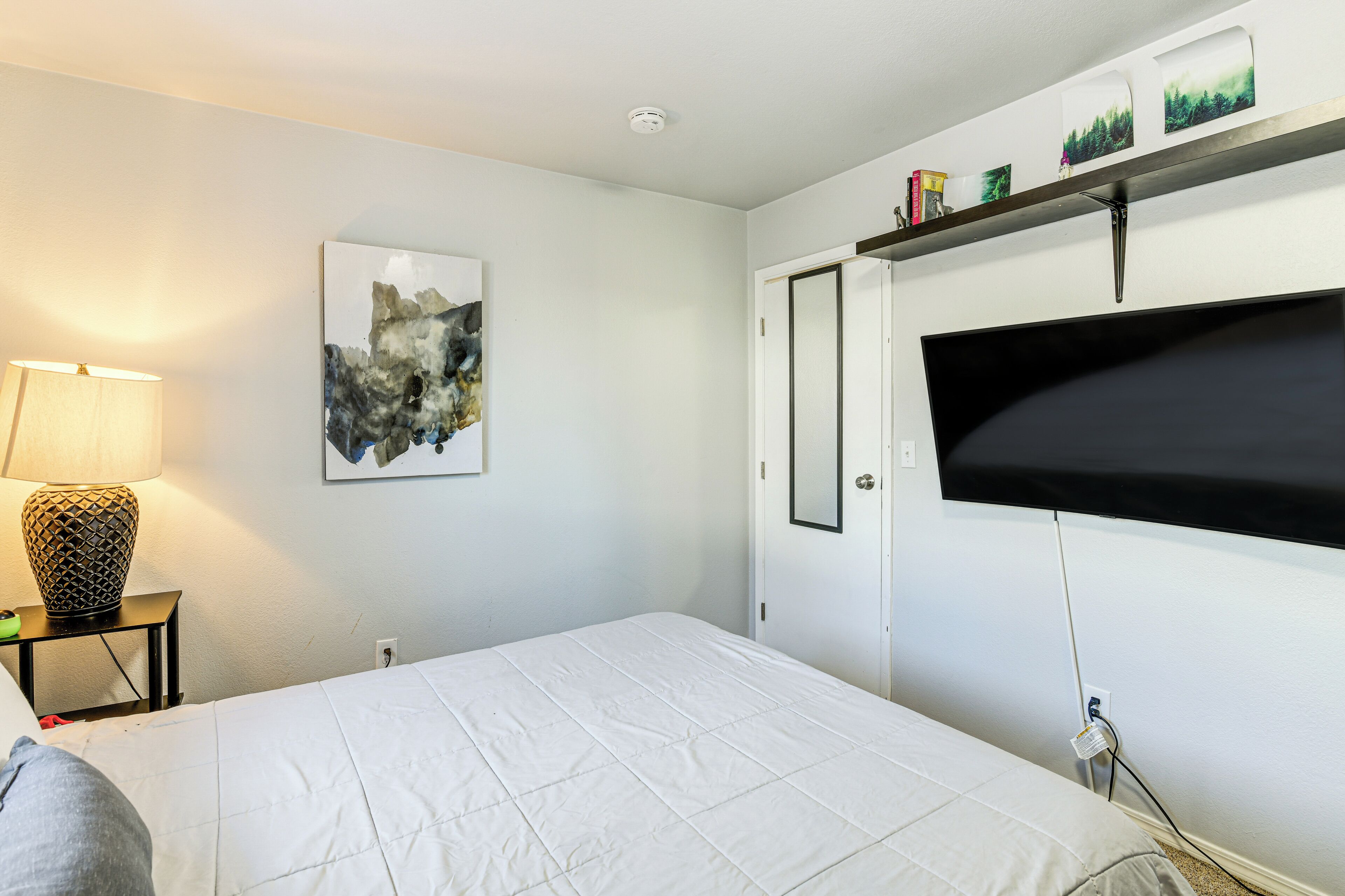 Een slaapkamer met een bed, een tv aan de muur, een schilderij en een plank met boeken en decoratieve objecten.