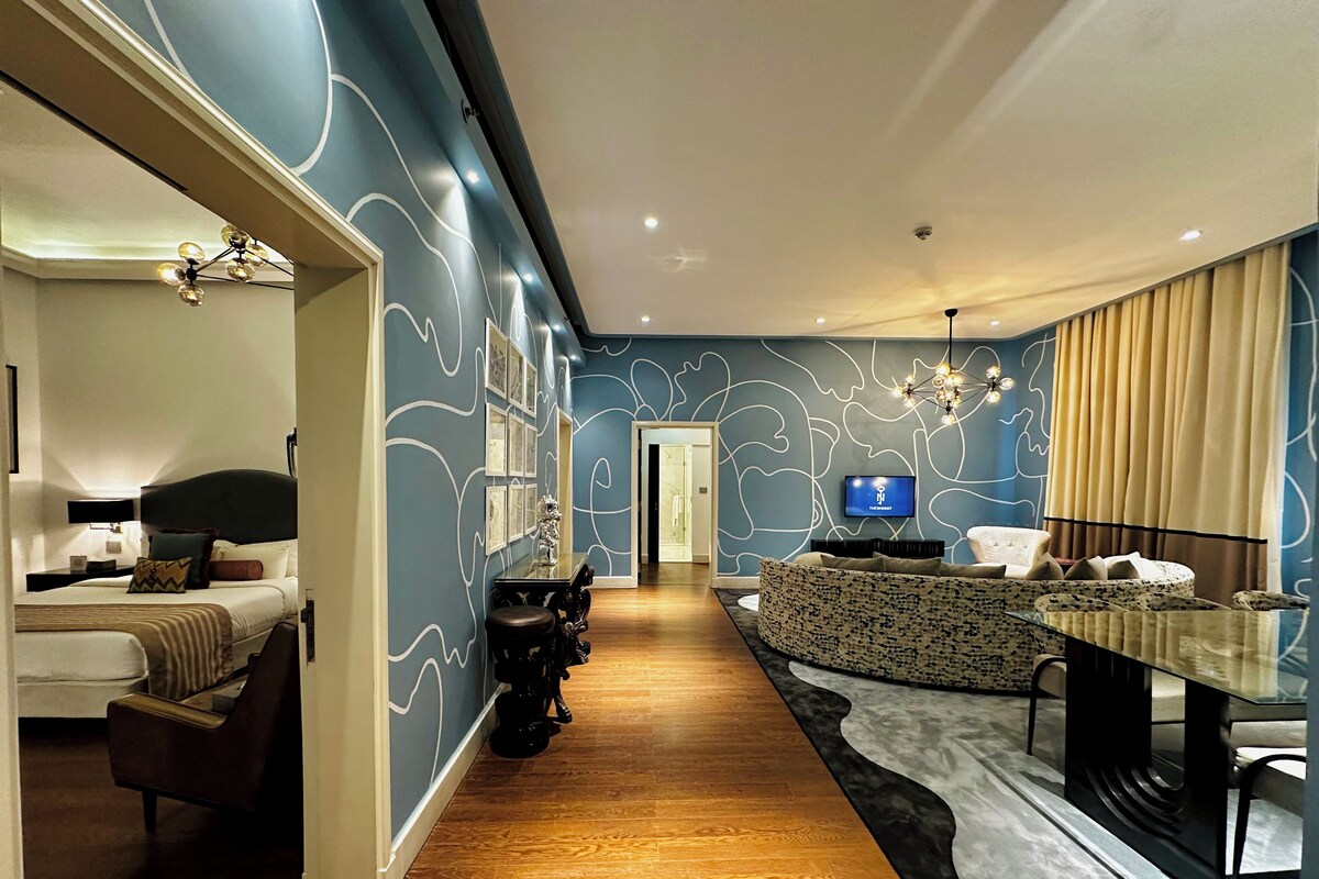 A modern hotel room with a bed, a seating area with a TV, and a hallway leading to another room.