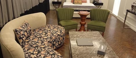 Une chambre d’hôtel avec un canapé, deux fauteuils verts, une table basse en marbre et un lit avec une tête de lit.