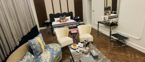 A modern hotel room with a large L-shaped sofa, a small round coffee table, a bed with a headboard, two armchairs, a vanity with a mirror, and a ceiling light fixture.