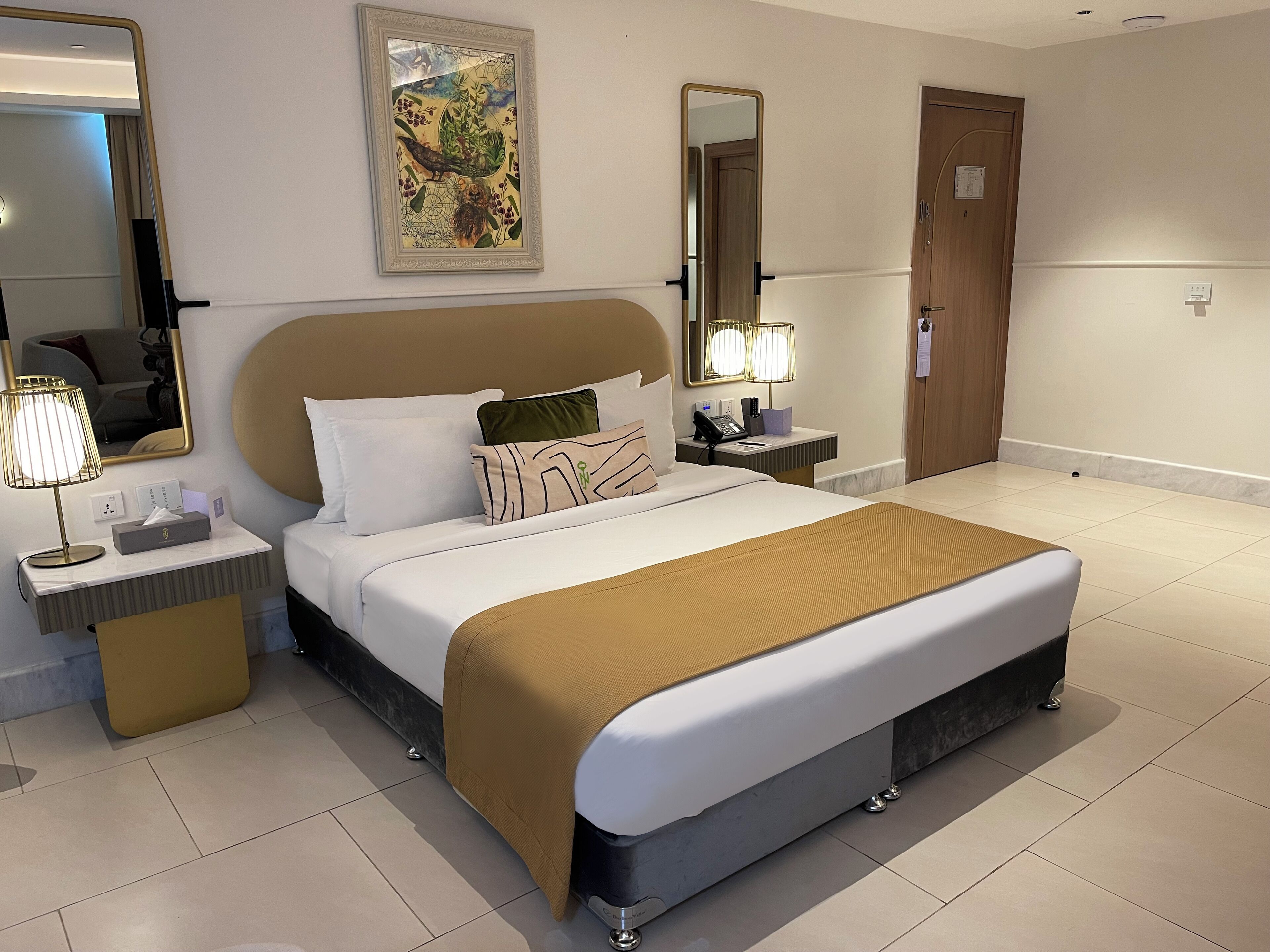 A hotel room with a large bed, two bedside tables, a nightstand, a mirror, and a painting on the wall.