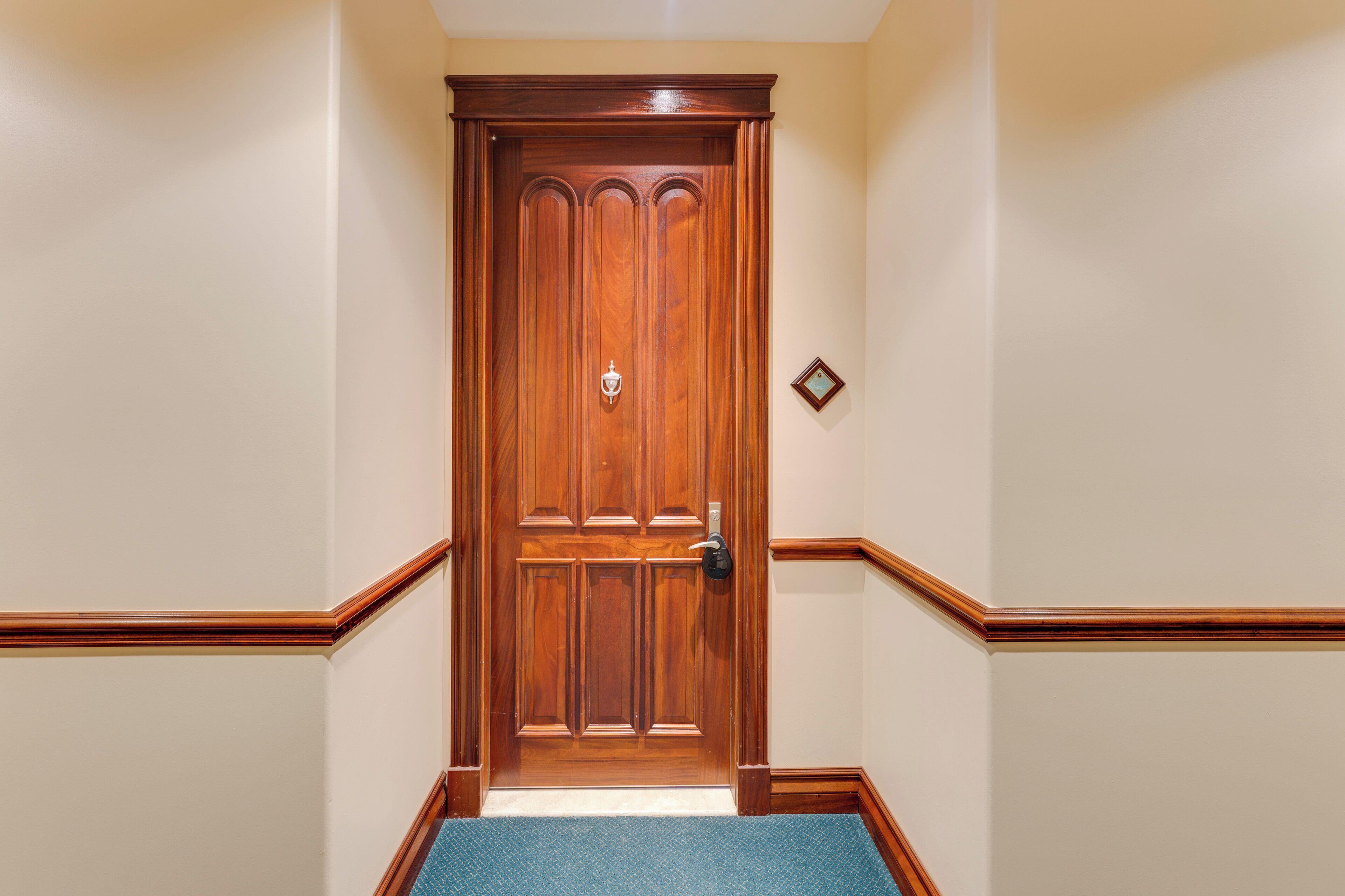 Puerta de madera con manija de latón y cerradura, enmarcada por paredes beige con detalles de madera.
