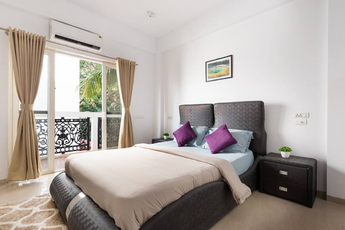 A bedroom with a large bed, a nightstand, a small table with a plant, and a window with curtains.