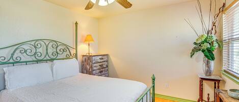 A bedroom with a bed, a ceiling fan, a nightstand, a lamp, a dresser, a window with blinds, and a plant.