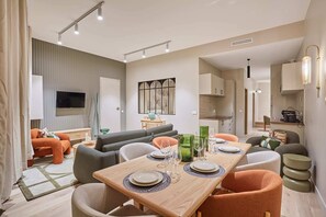 A modern living room with a dining area, grey sofas, orange armchairs, and a flat-screen TV mounted on the wall.