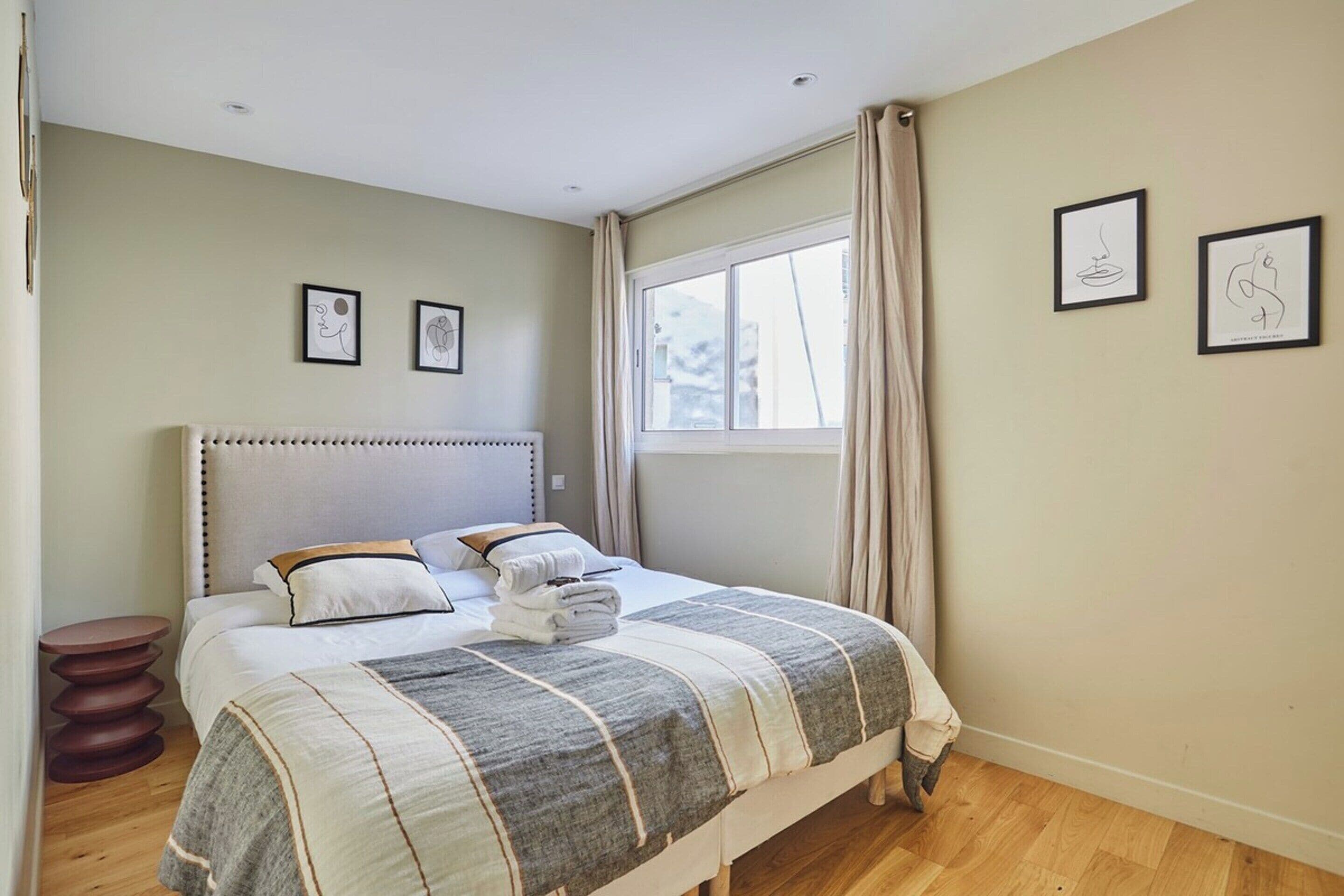 Een slaapkamer met een groot bed, een rond houten nachtkastje en twee kunstwerken in lijsten aan de muur.