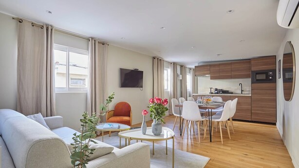 Ein modernes Wohnzimmer mit einer Couch, einem Esstisch und Stühlen, einer Küchenzeile mit Mikrowelle und Schränken sowie einem an der Wand montierten Fernseher.
