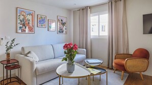 Ein modernes Wohnzimmer mit einer Couch, einem Couchtisch und einem Sessel. An den Wänden hängen gerahmte Kunstwerke und ein Fenster mit Vorhängen.