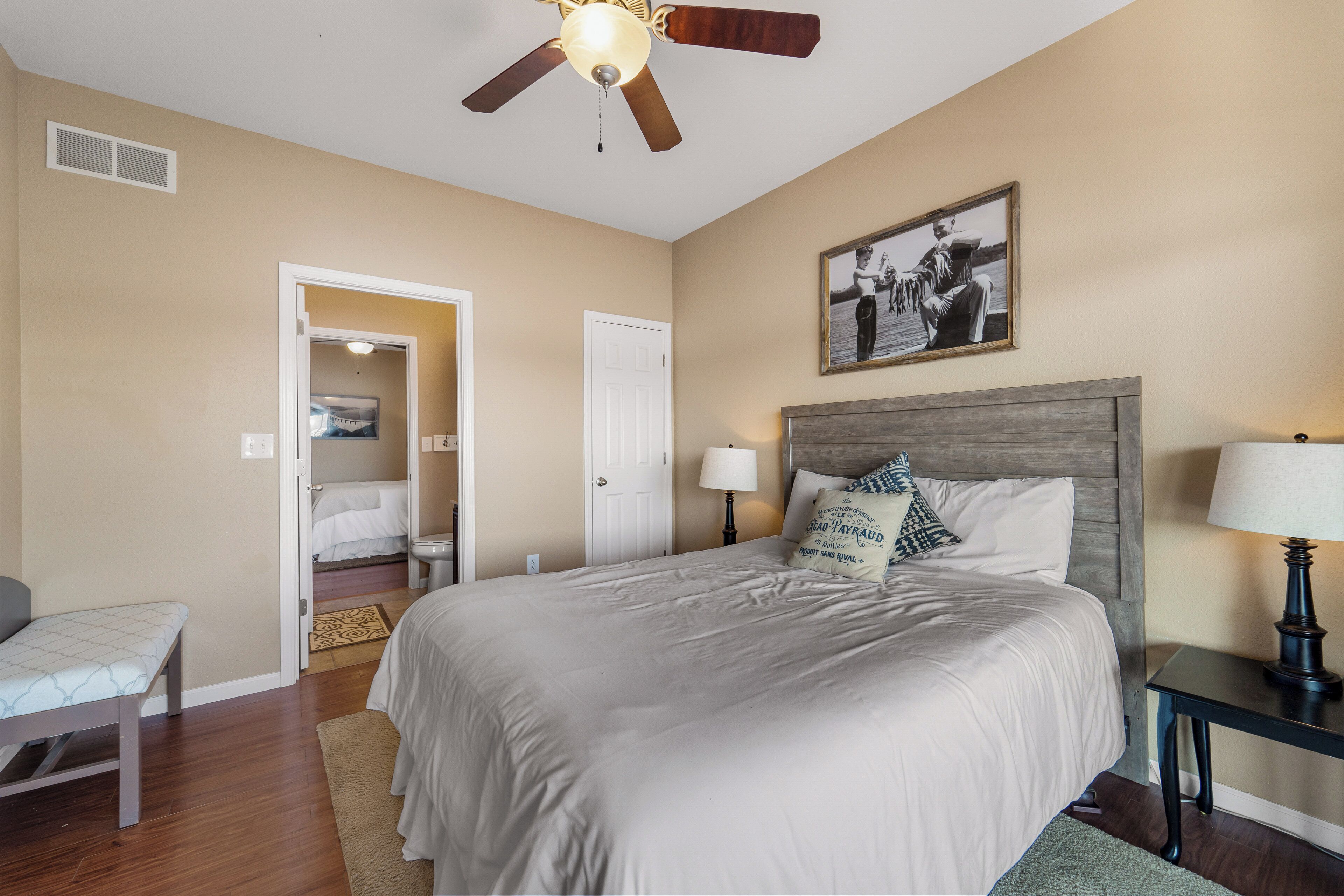 A bedroom with a large bed, a ceiling fan, a nightstand with a lamp, a bench, and a door leading to another room.