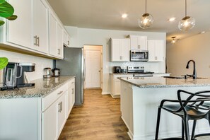 A modern kitchen with white cabinets, granite countertops, a stainless steel refrigerator, a microwave, and a coffee maker. There are bar stools and pendant lights. - Modern Townhome w/ Private Yard in Kernersville! (Kernersville)