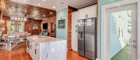 A modern kitchen with wooden ceiling, a marble island, stainless steel appliances, and a view through a glass door.