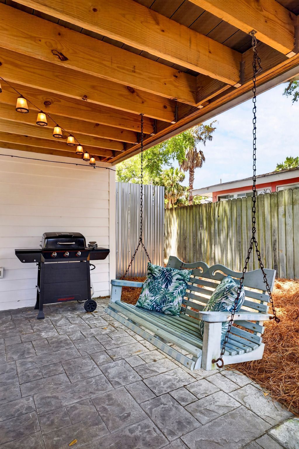 Een overdekt terras met een barbecue, een schommel en een vloer van stenen tegels.