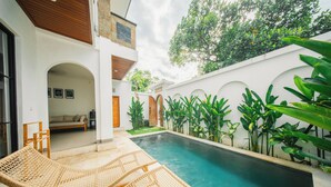 A pool area with two wicker lounge chairs, a small garden with tropical plants, and a white building with arched doorways. - 2BR Villa Seminyak with Pool (Seminyak, Badung Regency)