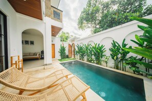 A pool area with two wicker lounge chairs, a small garden with tropical plants, and a white building with arched doorways. - 2BR Villa Seminyak with Pool (Seminyak, Badung Regency)