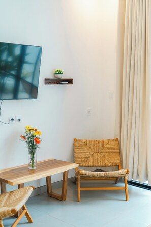 A modern living room with a wooden table, a wicker chair, a small shelf with decorative items, and a flat-screen TV mounted on the wall. - 2BR Villa Seminyak with Pool (Seminyak, Badung Regency)
