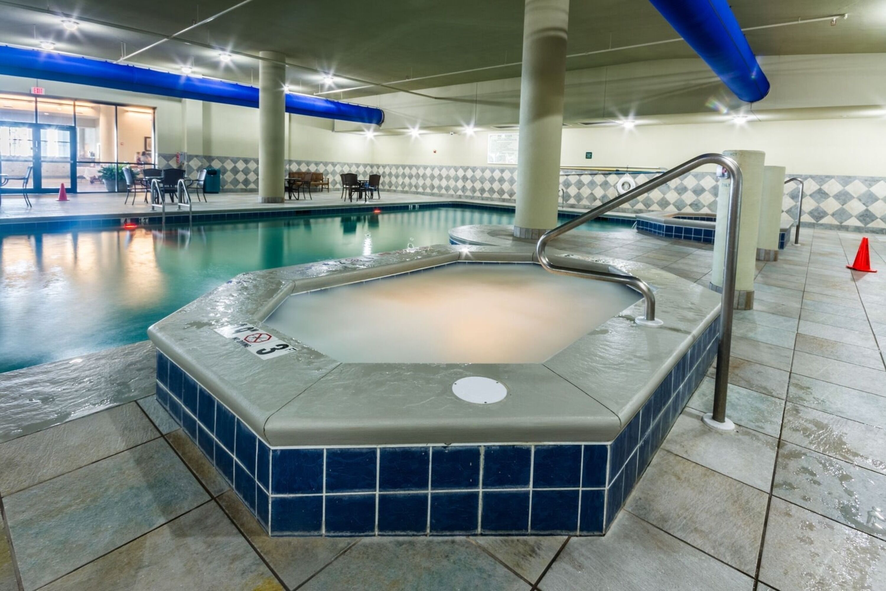 An indoor swimming pool with a hot tub, tiled walls, and a seating area with tables and chairs.