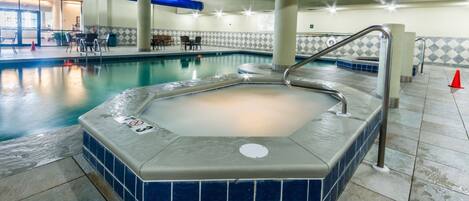 An indoor swimming pool with a hot tub, tiled walls, and a seating area with tables and chairs.