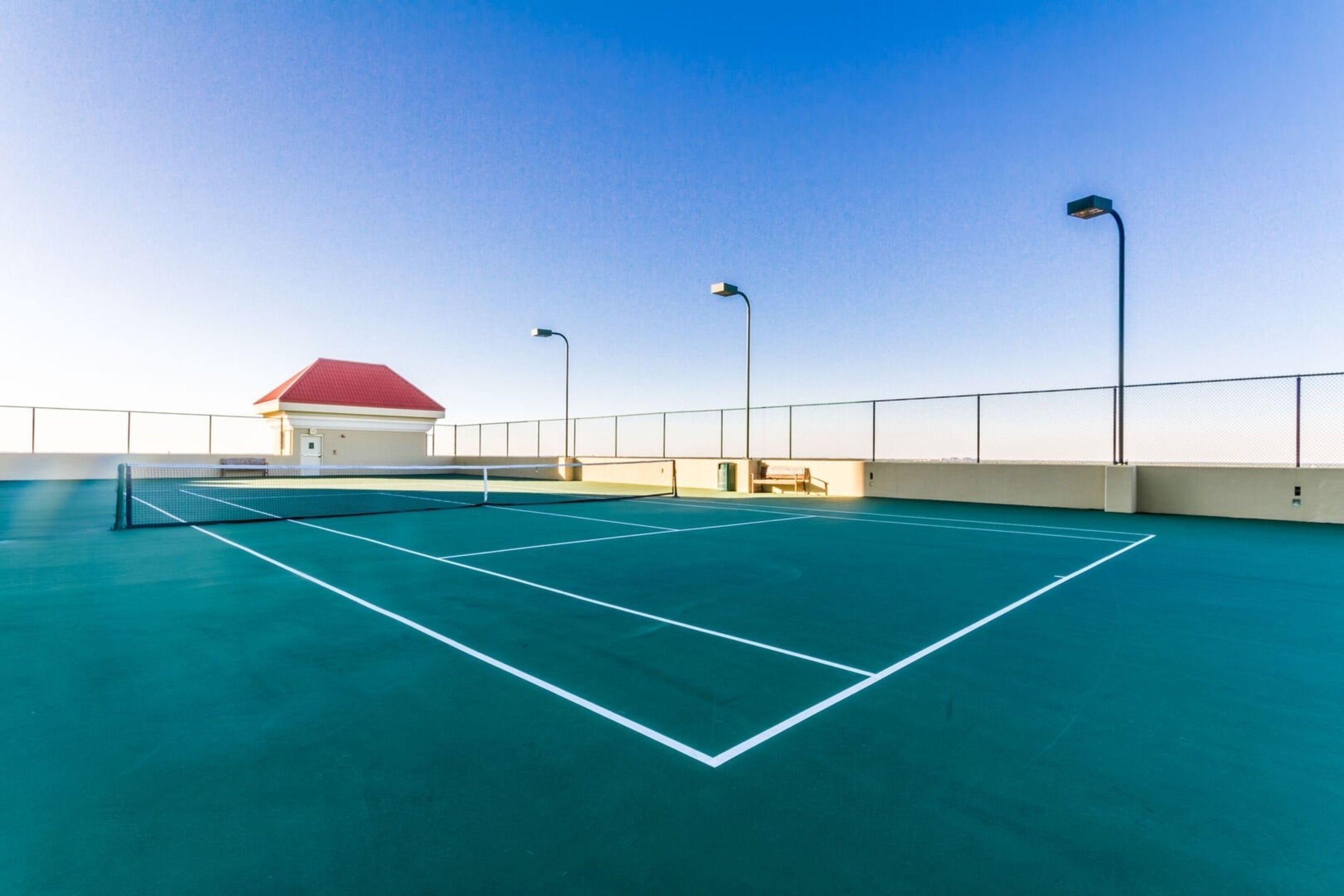 An indoor tennis court with a net, three street lamps, and a red-roofed building in the background.