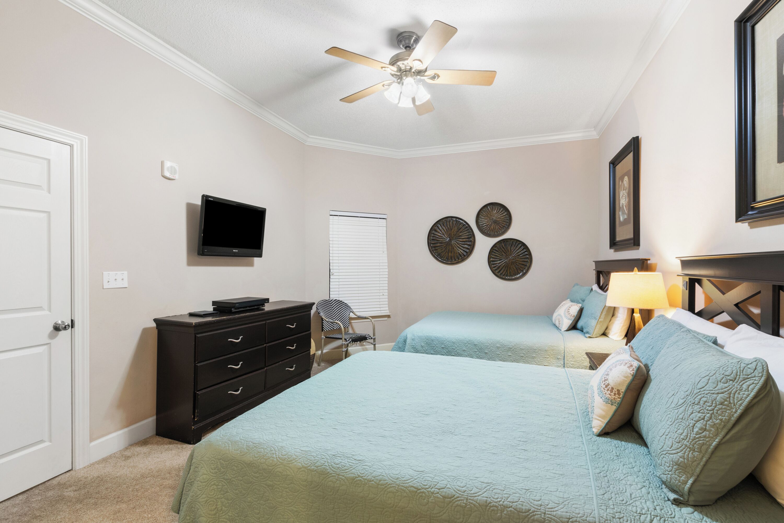 Une chambre avec deux lits, un ventilateur de plafond, une télévision, une commode et des œuvres d’art décoratives au mur.