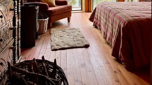 A cozy cabin room with a large bed, a wooden chair, a fireplace, and a view of the forest through large windows. - La Bella Patagonia (Cisnes)