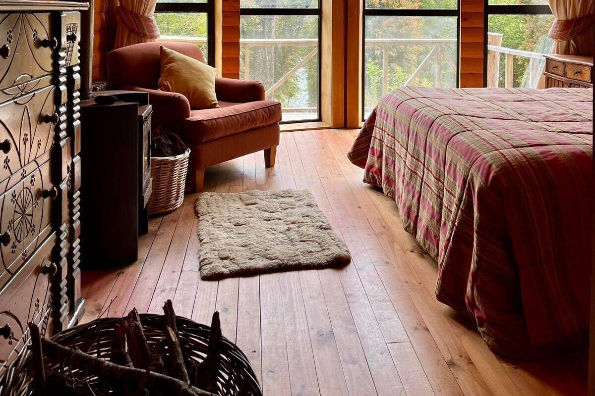 Quarto de cabana acolhedor com uma cama grande, uma cadeira de madeira, uma lareira e vista para a floresta através de amplas janelas.