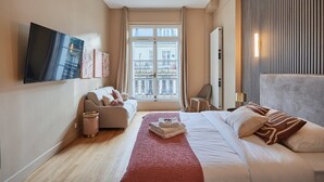 Ein modernes Hotelzimmer mit einem großen Bett, einer Couch, einem an der Wand befestigten Fernseher und einem Fenster mit Blick auf Gebäude.