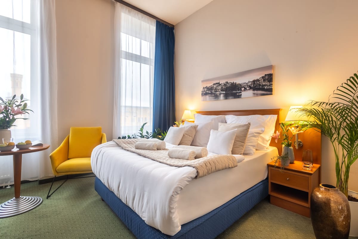 A hotel room with a large bed, a yellow armchair, a small table with a vase, and a large window with curtains.