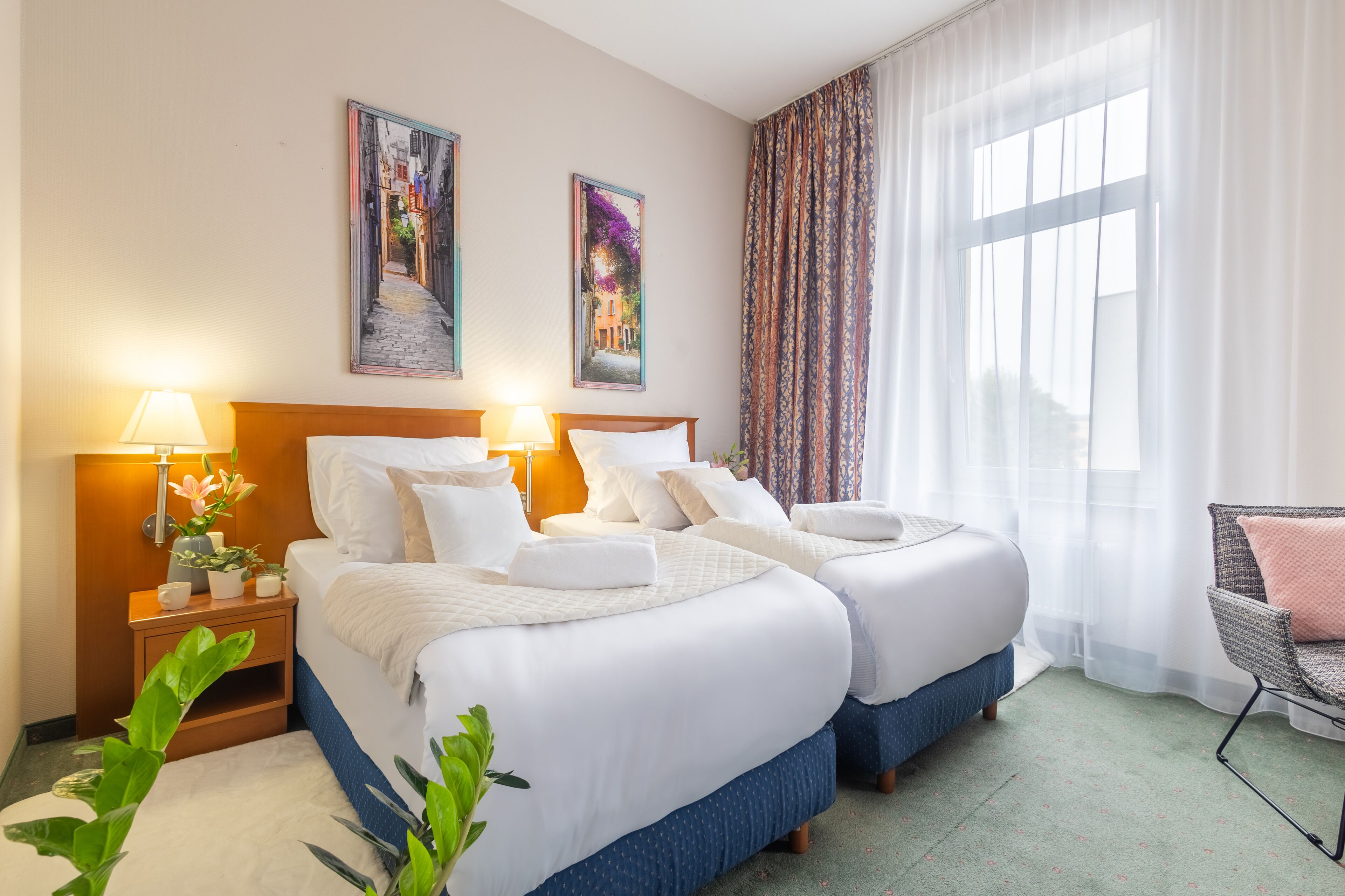 A hotel room with two beds, a nightstand with a lamp, a chair, and two framed pictures on the wall.
