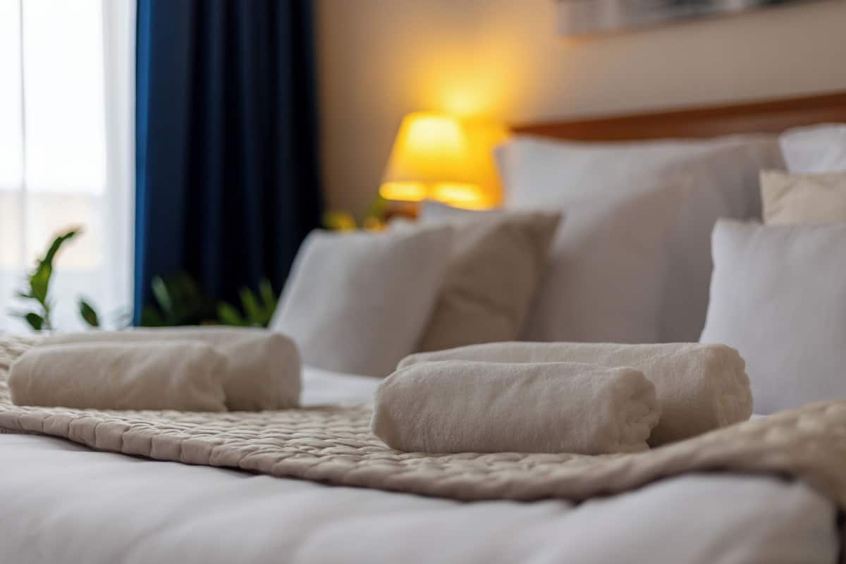 A neatly made bed with white linens and pillows, a textured blanket, and a bedside lamp in the background.