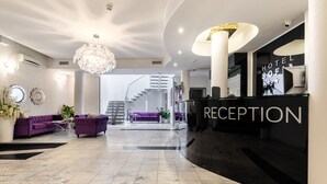 Un moderno mostrador de recepción de hotel con suelo de mármol, sofás morados, una araña y una escalera al fondo.