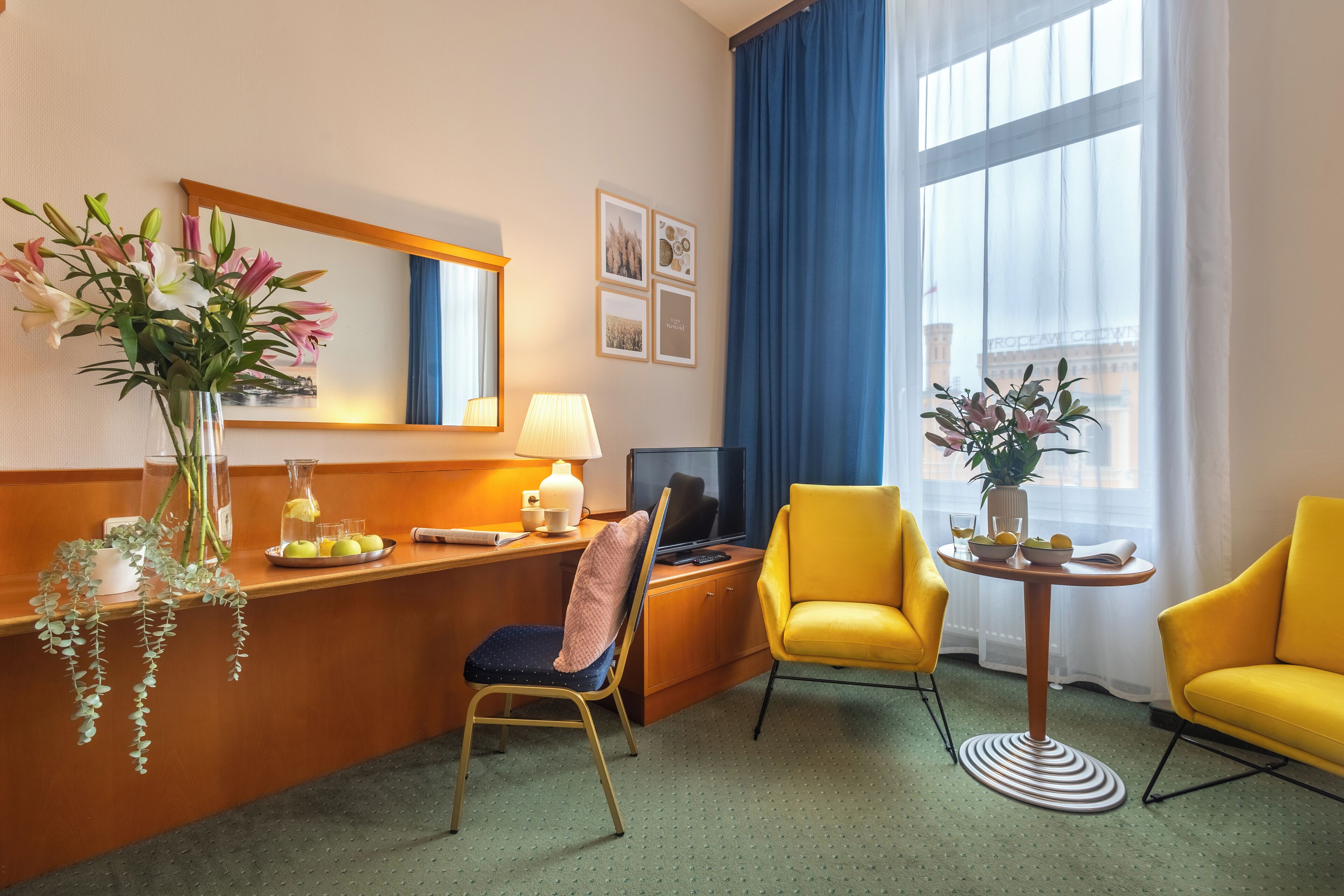 Een hotelkamer met een houten bureau, een televisie, een gele fauteuil, een klein rond tafeltje met een bloevas en een groot raam met blauwe gordijnen.