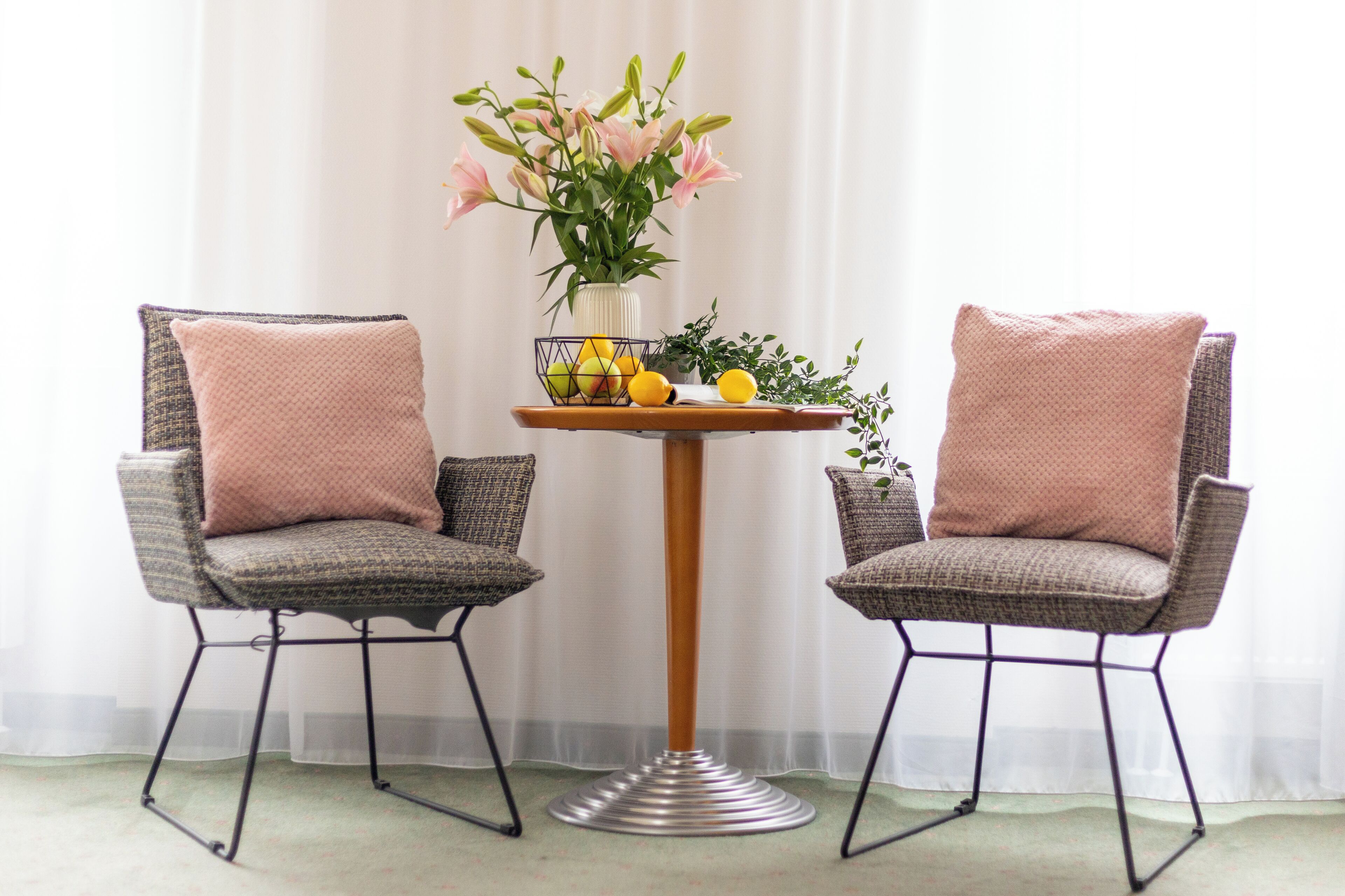 Two wicker chairs with cushions, a small round table with a vase of flowers and lemons, and a sheer white curtain in the background.