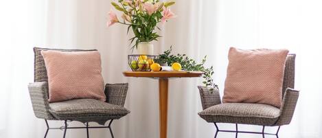 Two wicker chairs with cushions, a small round table with a vase of flowers and lemons, and a sheer white curtain in the background.