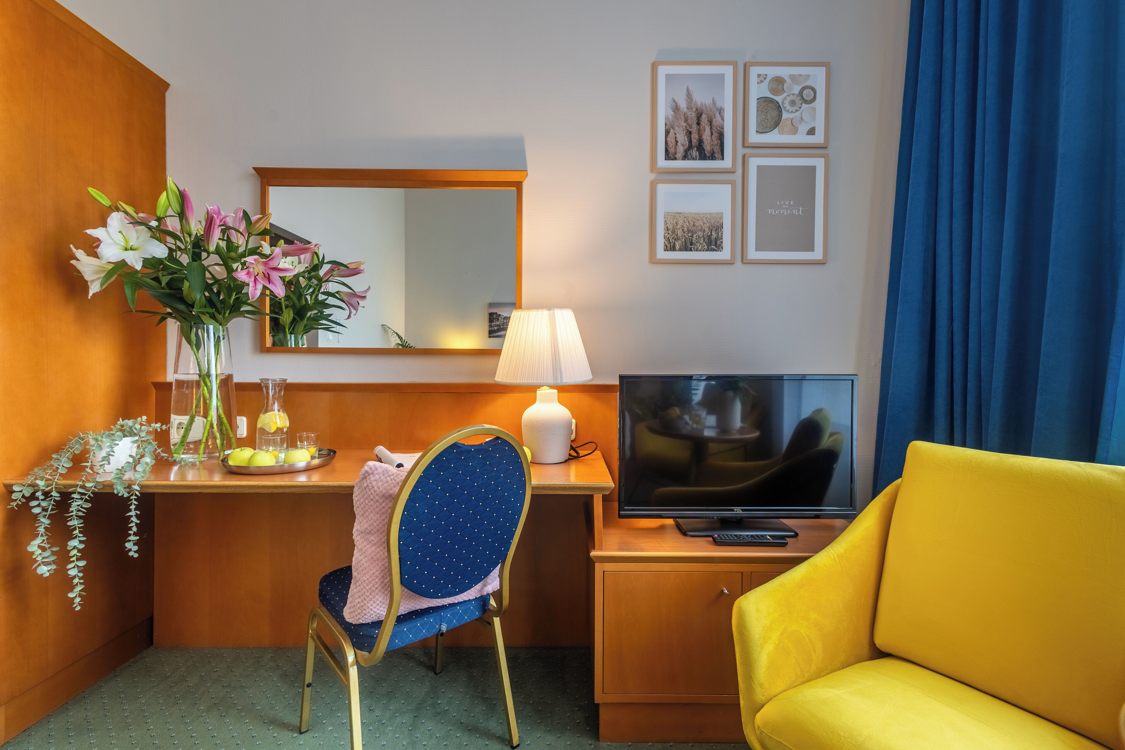 Una camera d'albergo con una scrivania in legno, una sedia, una televisione, una poltrona gialla, uno specchio e quadri appesi al muro.