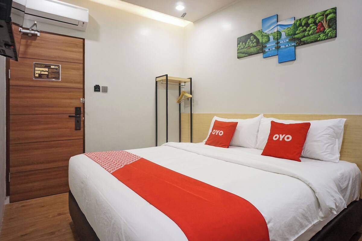 Une chambre d’hôtel avec une porte en bois, un lit avec une literie blanche et rouge, et une œuvre d’art accrochée au mur.