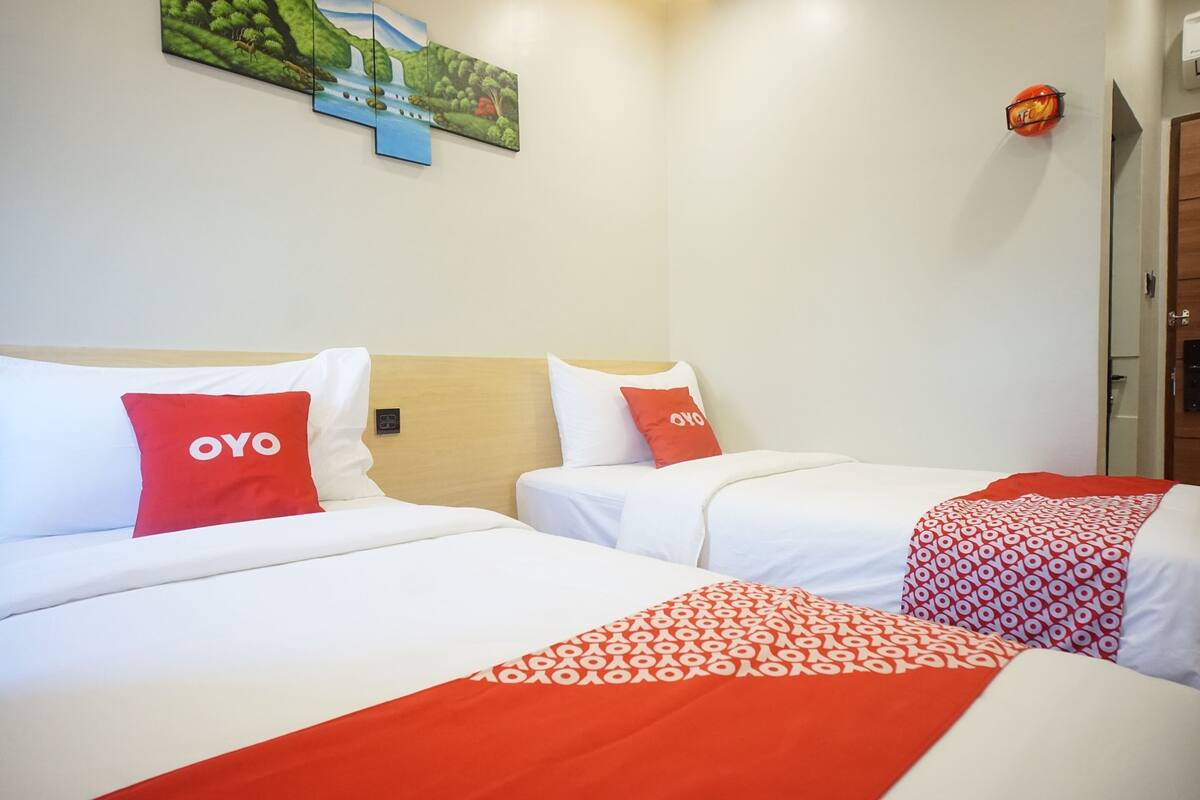 Une chambre d’hôtel avec deux lits, chacun étant équipé de draps blancs et d’oreillers rouges ornés du logo « OYO ». Un tableau est accroché au mur au-dessus des lits, et une porte se trouve sur le côté droit.