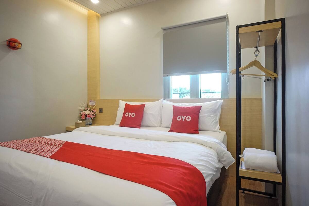 Une chambre d’hôtel avec un grand lit, deux oreillers rouges, une tête de lit en bois, une fenêtre avec vue, une table de chevet ornée d’un vase de fleurs et un porte-manteau mural.